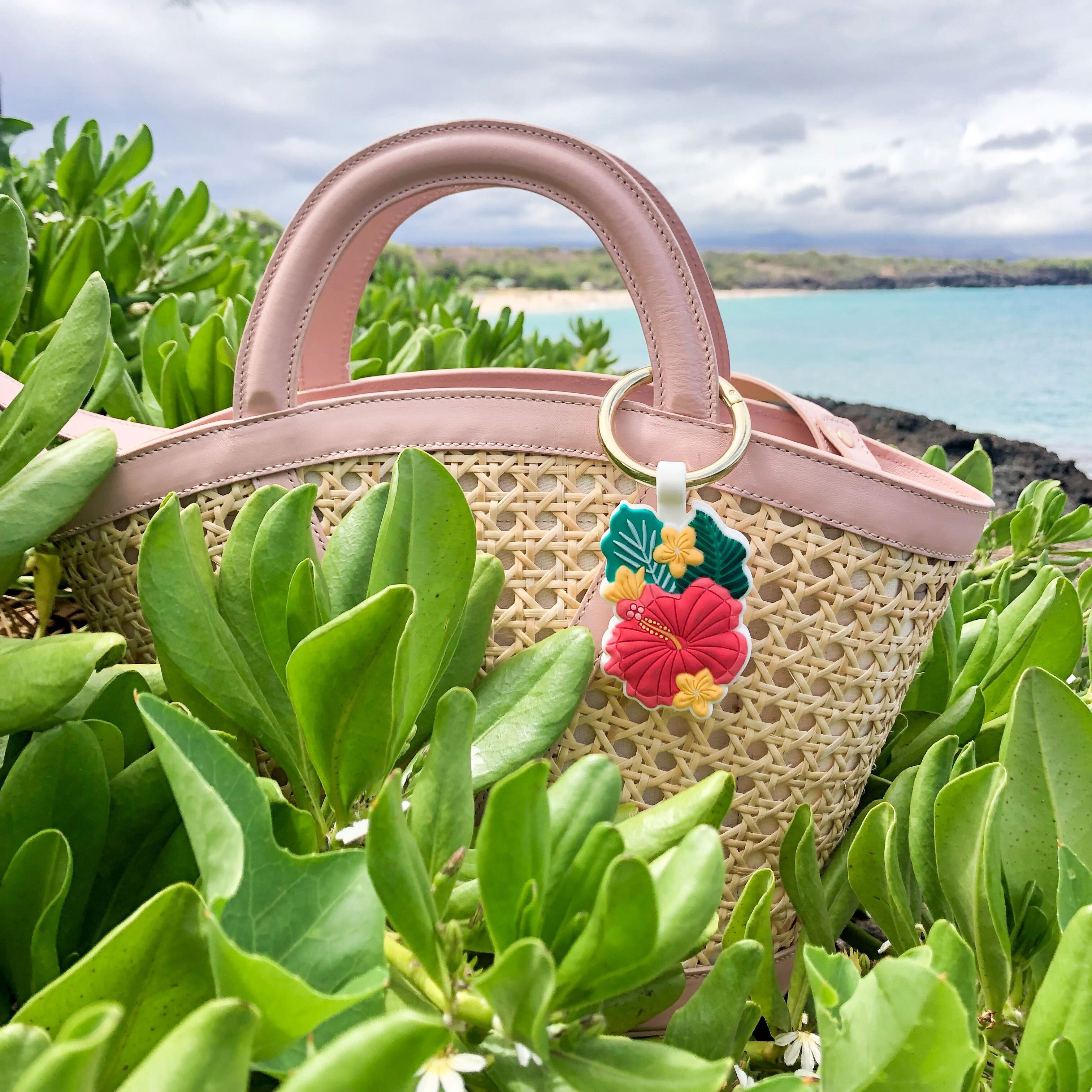 Maui, the limited-edition Bagnet magnetic purse holder, clipped to a rattan and leather basket bag, nesting in foliage with the blue Hawaiian coastline in the distance 