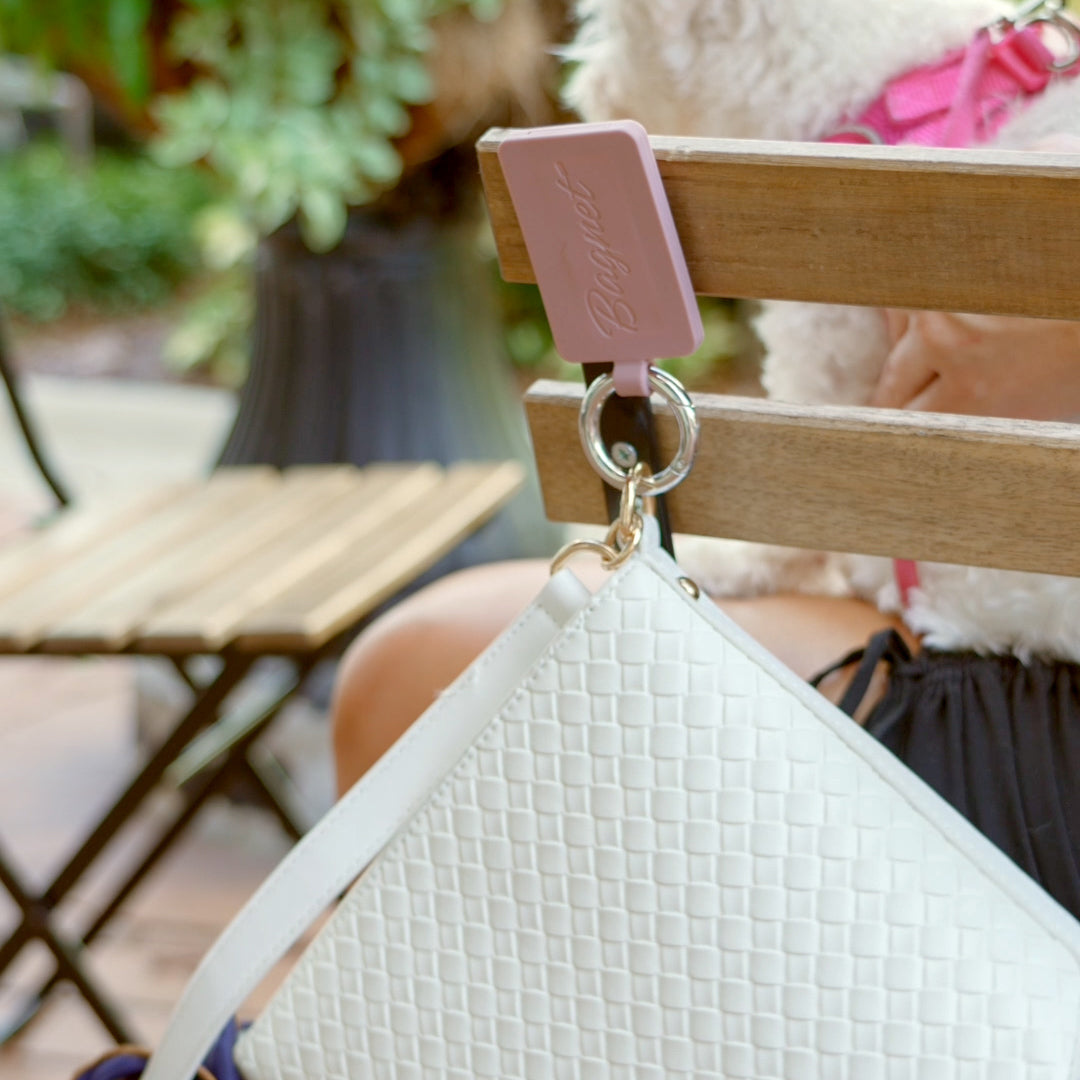 Burlwood Sport Bagnet holding a white purse, attached to the back of a metal chair in a green outdoor seating area