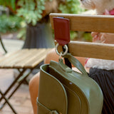 Merlot Leather Bagnet holding a green purse, attached to the back of a metal chair in a green outdoor seating area