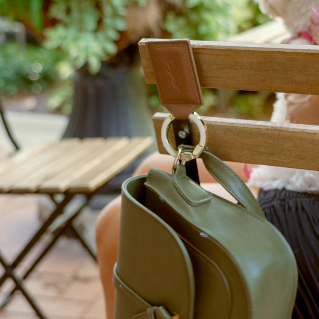 Saddle Leather Bagnet holding a green purse, attached to the back of a metal chair in a green outdoor seating area