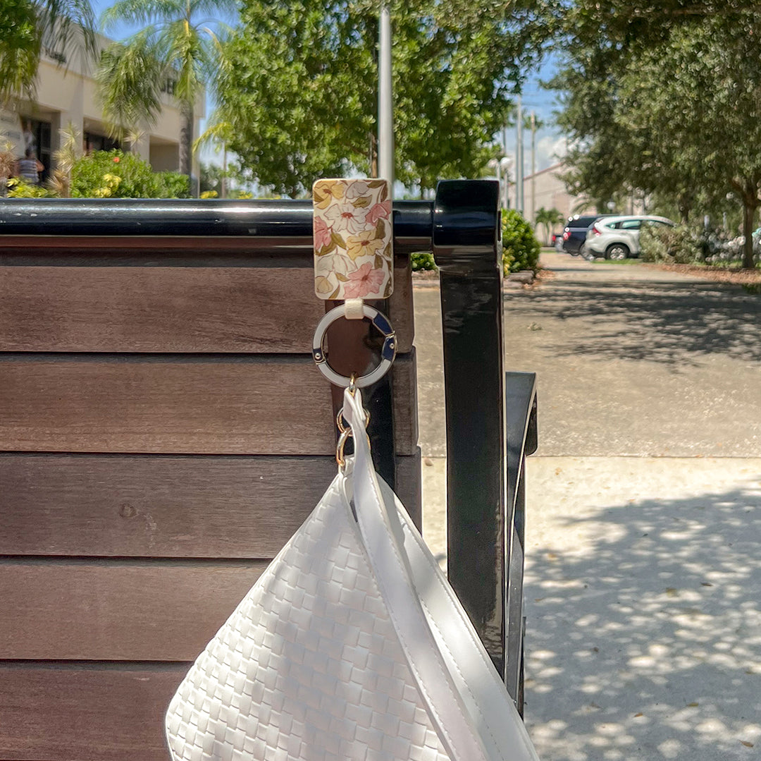 Pressed Petals Graphic Sport Bagnet holding a white purse, attached to the back of a metal bench in a green outdoor seating area