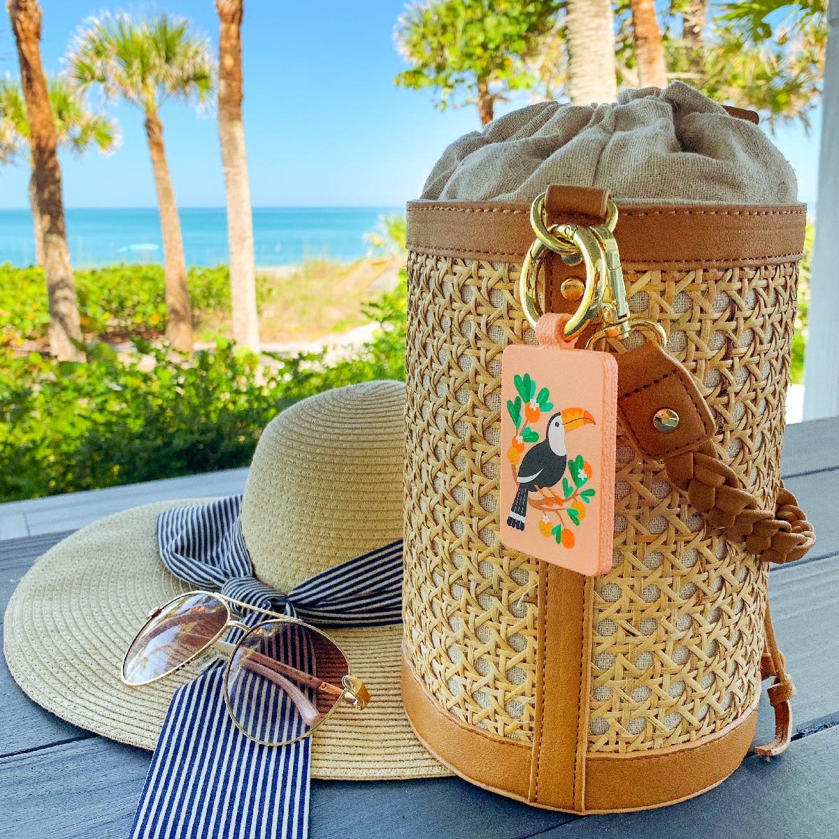Sammy, the limited-edition Bagnet magnetic purse holder, clipped to a rattan and leather basket bag, sitting on a deck with the blue coastline in the distance